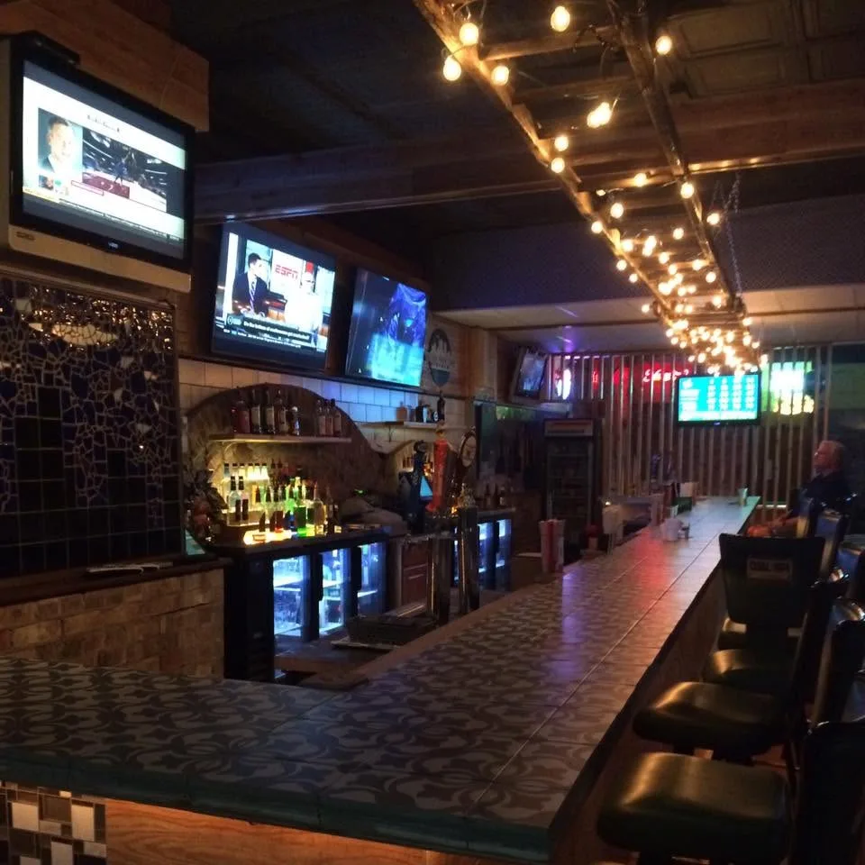 The long tiled bar under strings of lights, stools along it and televisions above